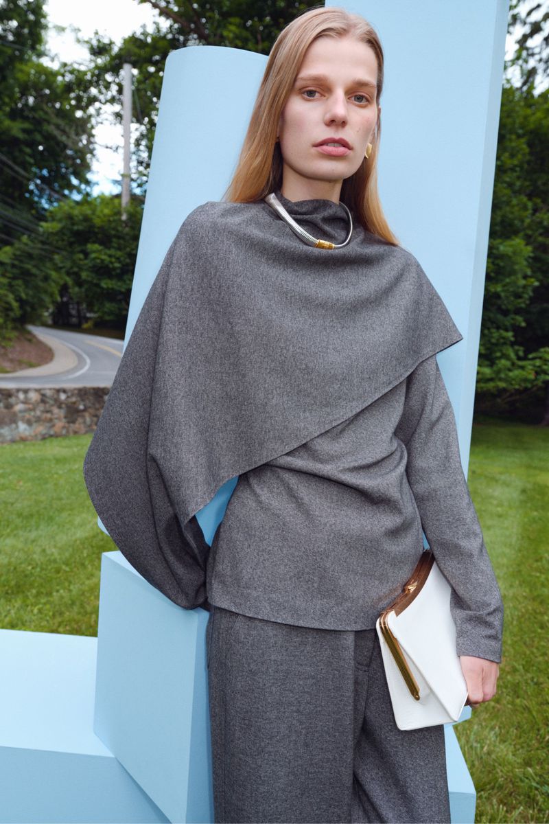 Model wearing a draped grey cape sweater and tailored pants holding a white clutch against light blue panels outdoors.
