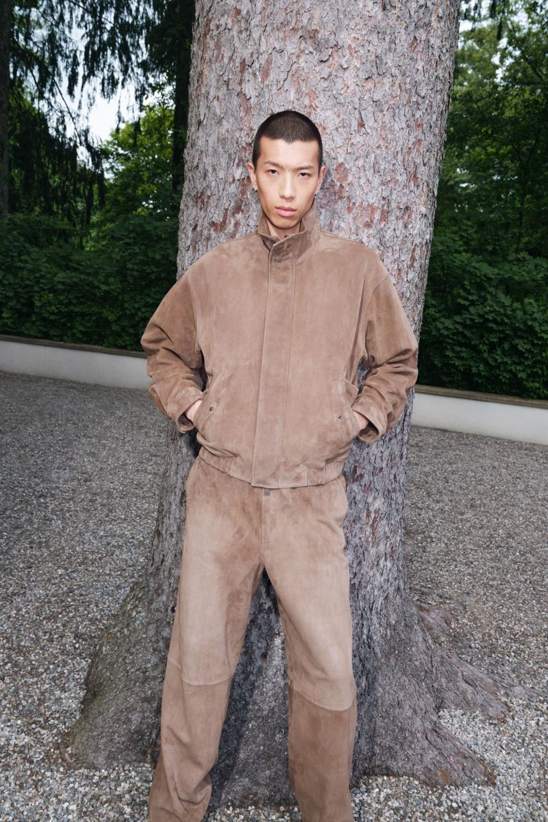 Model Cai Yuyang wearing a tan suede jacket and matching trousers against a tree trunk for Simkhai.
