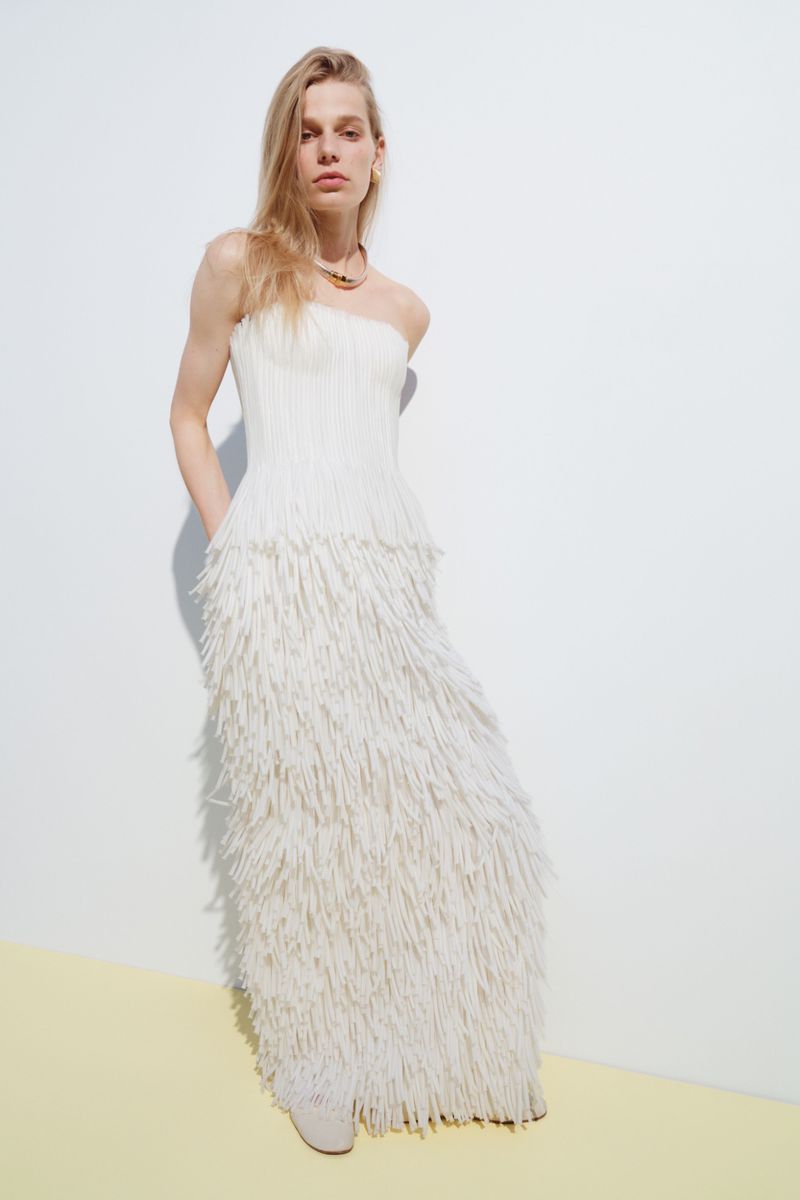 Model wearing a strapless off-white fringe maxi dress and a gold choker necklace against a light studio backdrop.