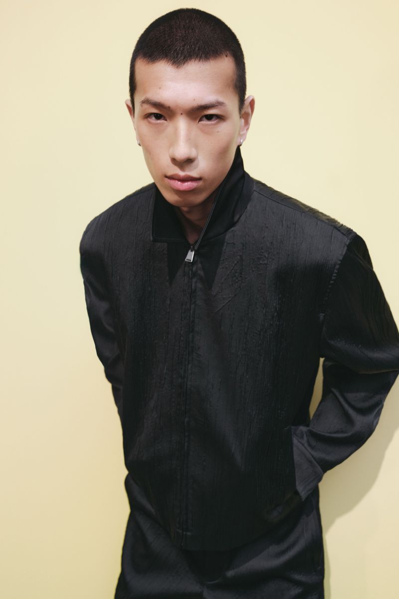 East Asian male model with short hair wearing a textured black zip jacket against a pale yellow seamless backdrop.