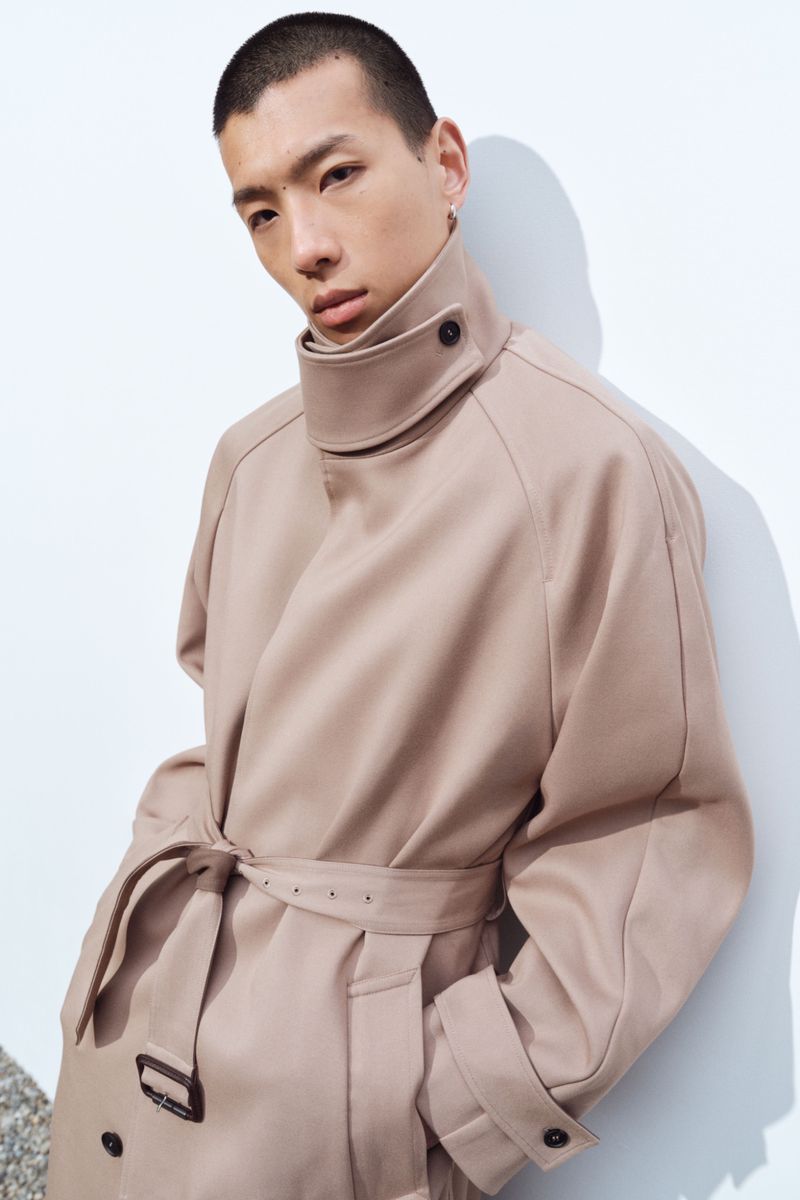 Model Cai Yuyang wearing an oversized beige high-collar belted trench coat against a white wall for the Simkhai Resort lookbook.