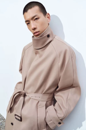 Model Cai Yuyang wearing an oversized beige high-collar belted trench coat against a white wall for the Simkhai Resort lookbook.