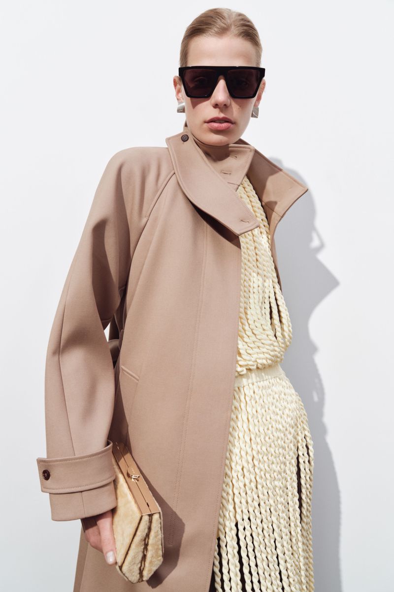 Model wearing dark flat-top sunglasses, a tailored beige coat, and a cream rope-fringe knit dress against a white background.