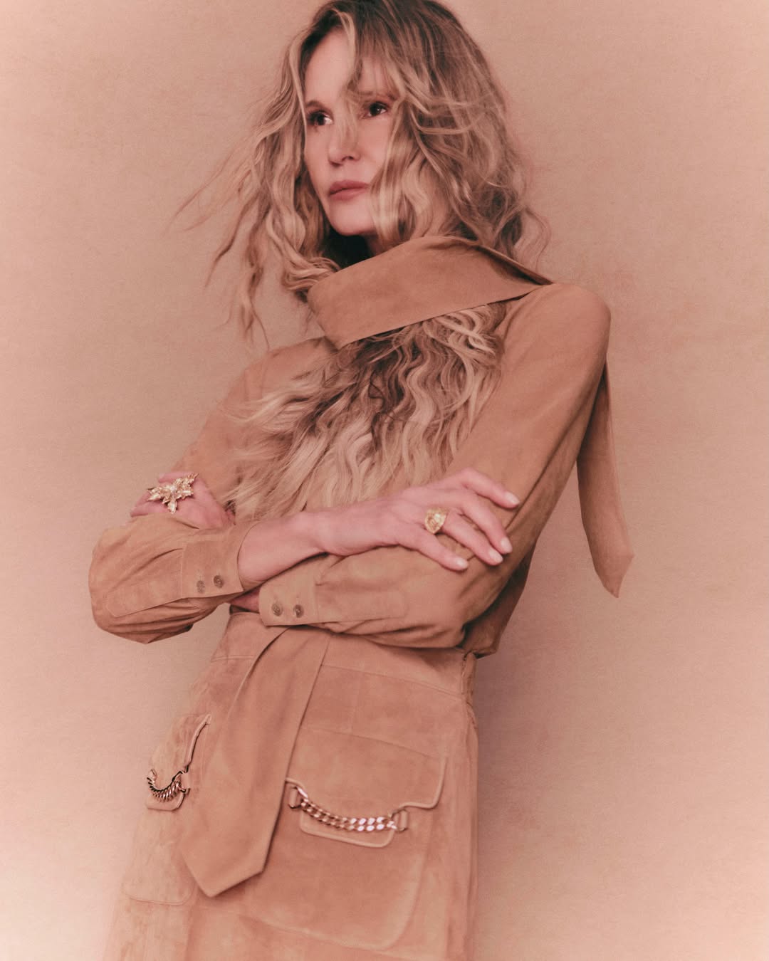 Model Elle Macpherson posing with crossed arms in a tan suede dress with a neck wrap detail against a warm beige studio background.