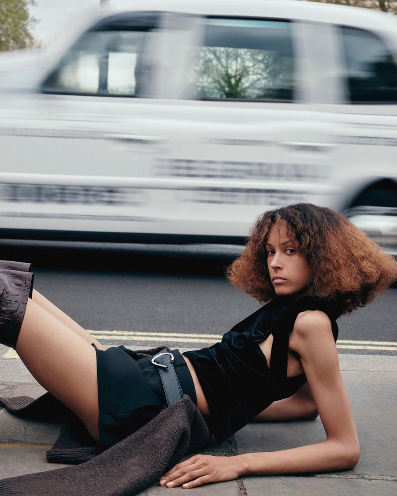 Woman with curly hair in a black top and shorts lying on a city sidewalk next to a blurry white cab and trees in the background.