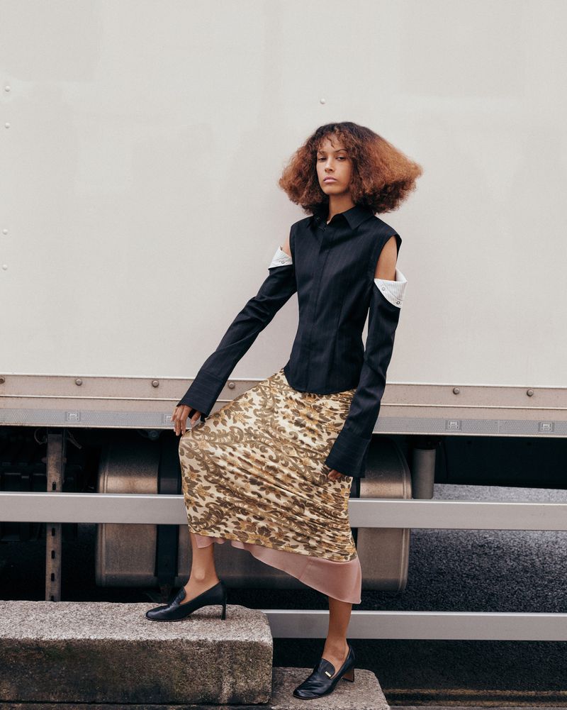 A stylish fashion portrait of a curly-haired woman in a black shirt and animal-print skirt, posing outdoors on concrete steps by a white truck.