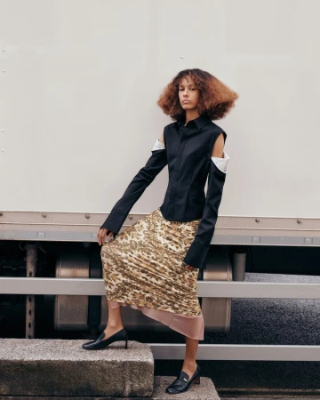A stylish fashion portrait of a curly-haired woman in a black shirt and animal-print skirt, posing outdoors on concrete steps by a white truck.
