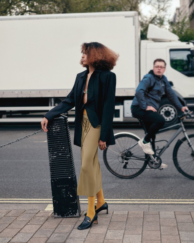 Woman in blazer and skirt on city street with cyclist and truck in background; fashion and urban photography on a cloudy day.