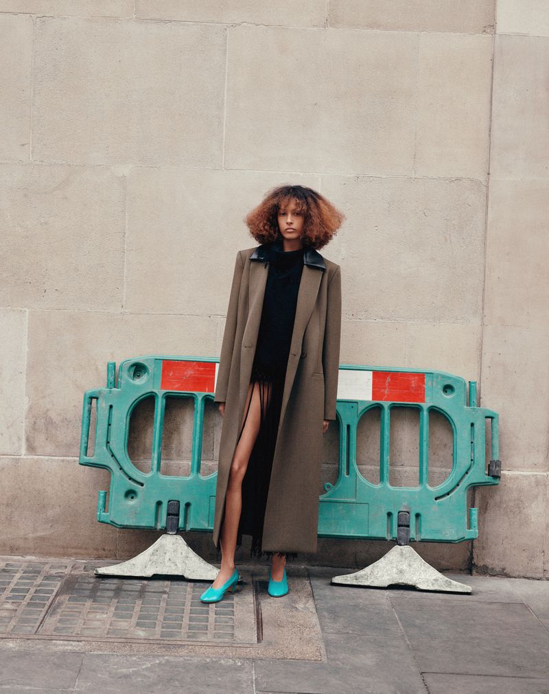 Woman in a long coat poses in front of a wall and a green barrier. She wears blue shoes and has curly auburn hair.