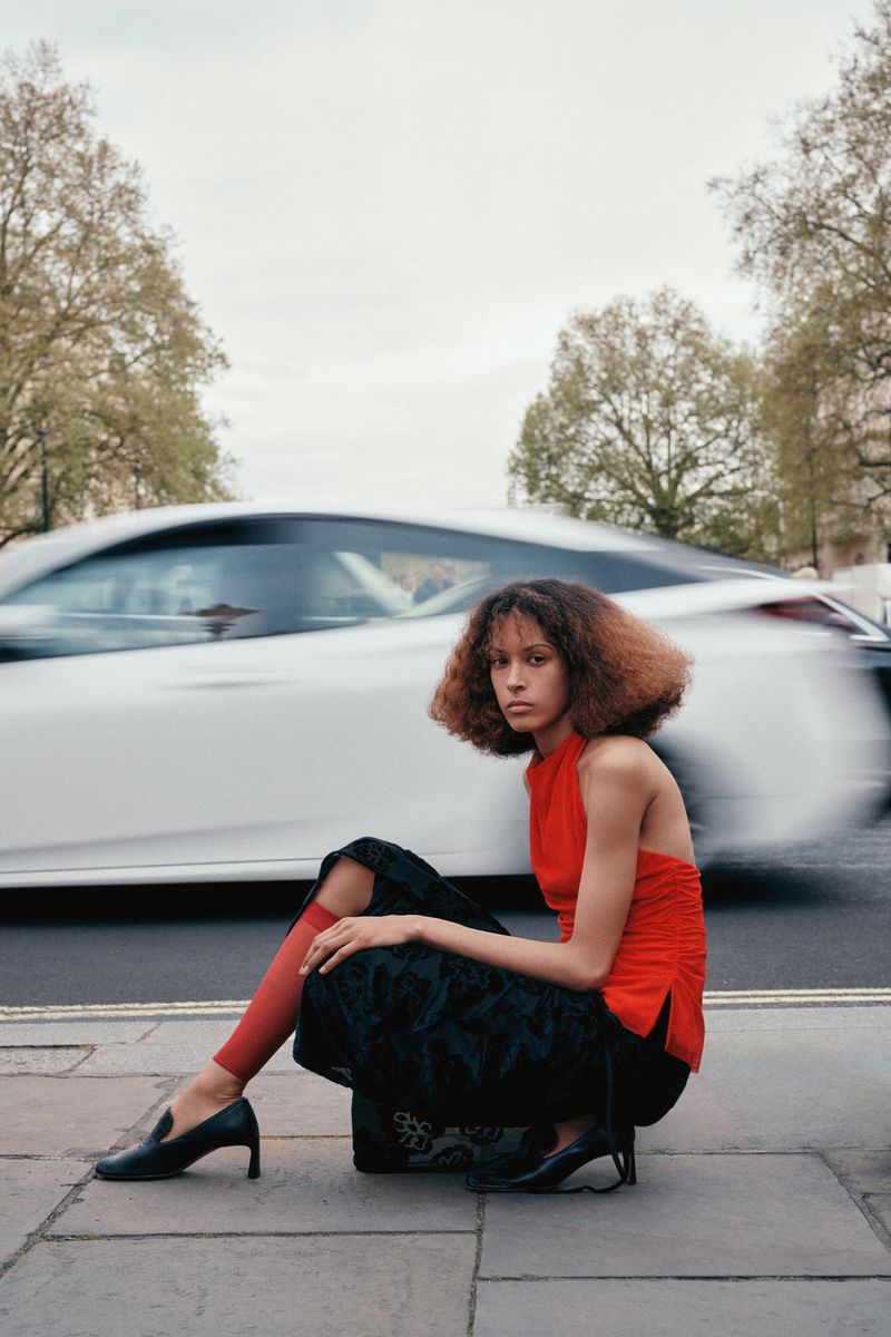 Fashion shot of a woman in an orange top and black skirt crouching outdoors, with a blurred white car passing in the background.