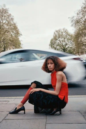 Fashion shot of a woman in an orange top and black skirt crouching outdoors, with a blurred white car passing in the background.