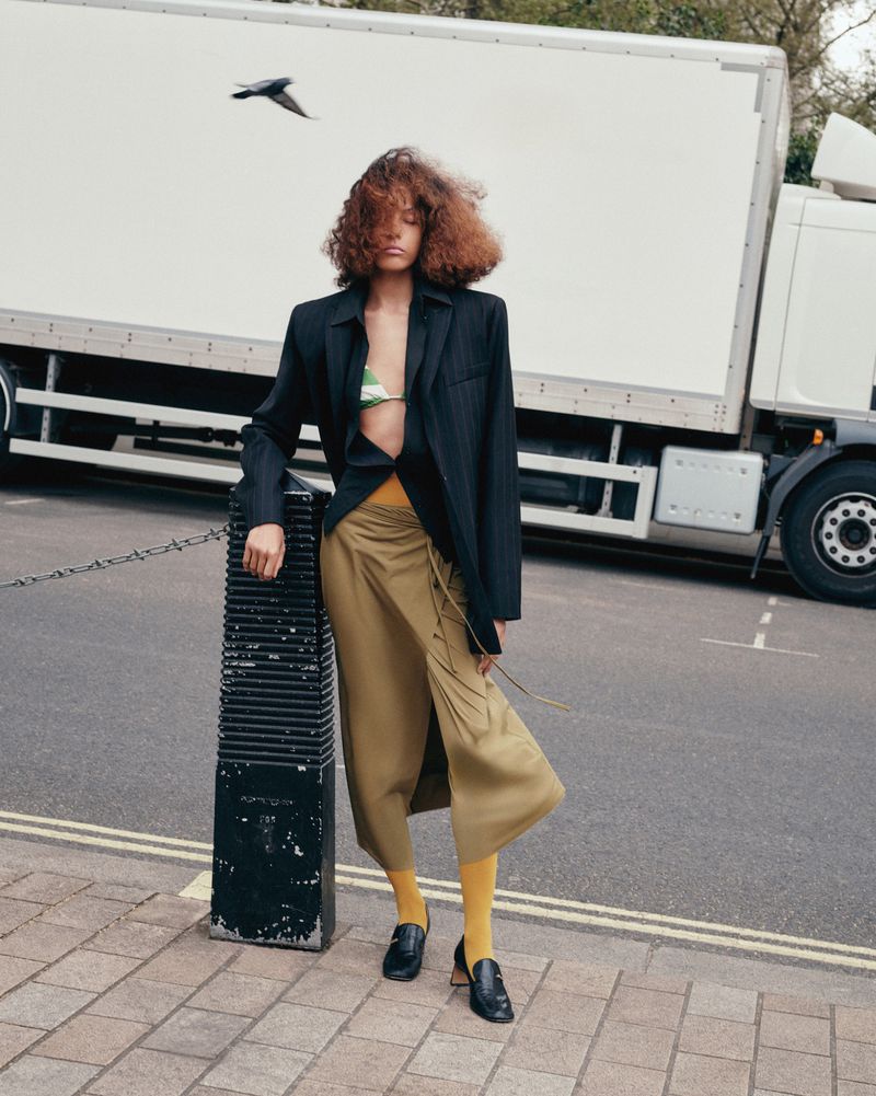 A woman with curly hair stands in a pinstripe blazer, draped khaki skirt, yellow tights, and black loafers, posing on an urban street.