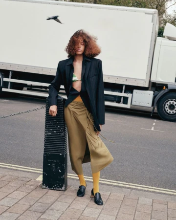 A woman with curly hair stands in a pinstripe blazer, draped khaki skirt, yellow tights, and black loafers, posing on an urban street.