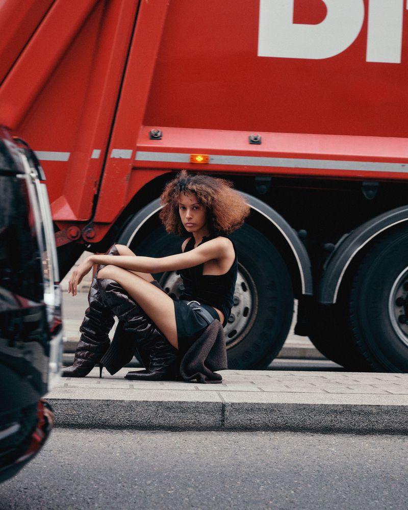 Young woman poses in tall, dark boots and a black top, seated on a curb next to a large red truck with a glimpse of a black car nearby.