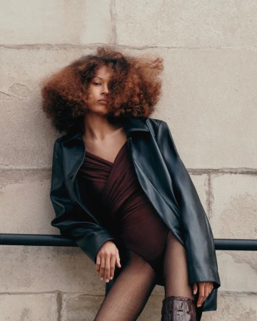 A woman with curly auburn hair poses outdoors wearing a black leather coat, burgundy bodysuit, tights, and knee-high boots for a fashion shot.