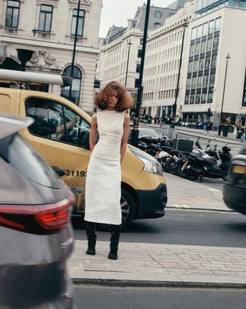 Woman in white dress stands on city street with yellow van and motorbikes in background, fashion shoot, outdoor daytime scene.