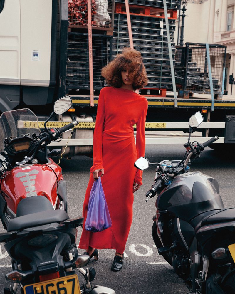 A woman in a long red dress stands between two motorcycles on a city street, holding a purple bag with a truck in the background.