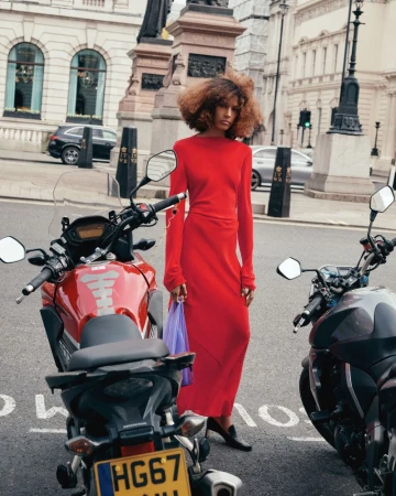 Prinnie Stott wearing a long red dress and black heels standing next to parked motorcycles on a city street.