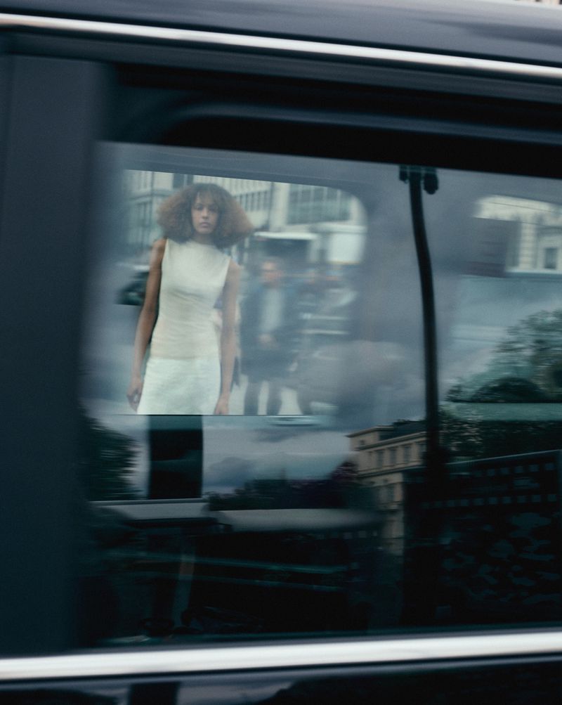 Fashion shot through a car window features an adult female model in an ivory dress standing on a city street with blurred figures in the background.