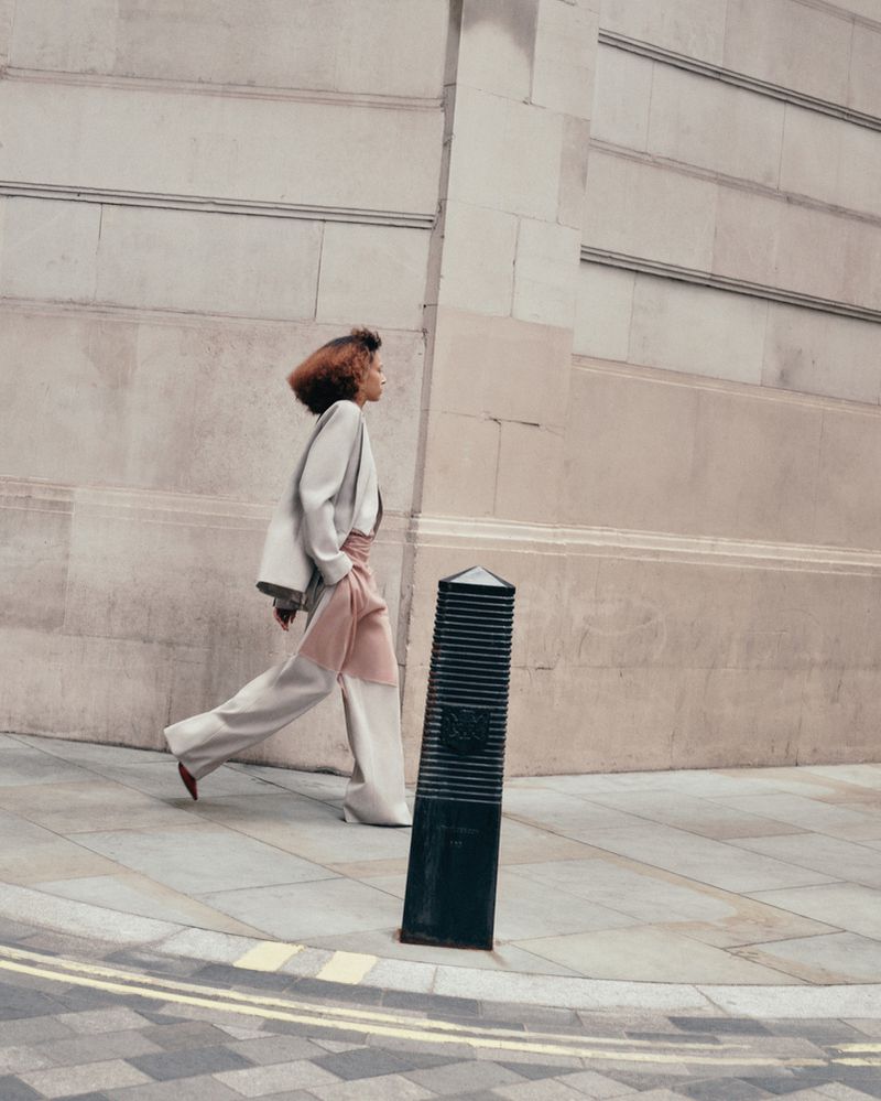 A fashion model in an oversized blazer and two-tone pants walks on a sidewalk next to a beige stone wall with a black bollard.