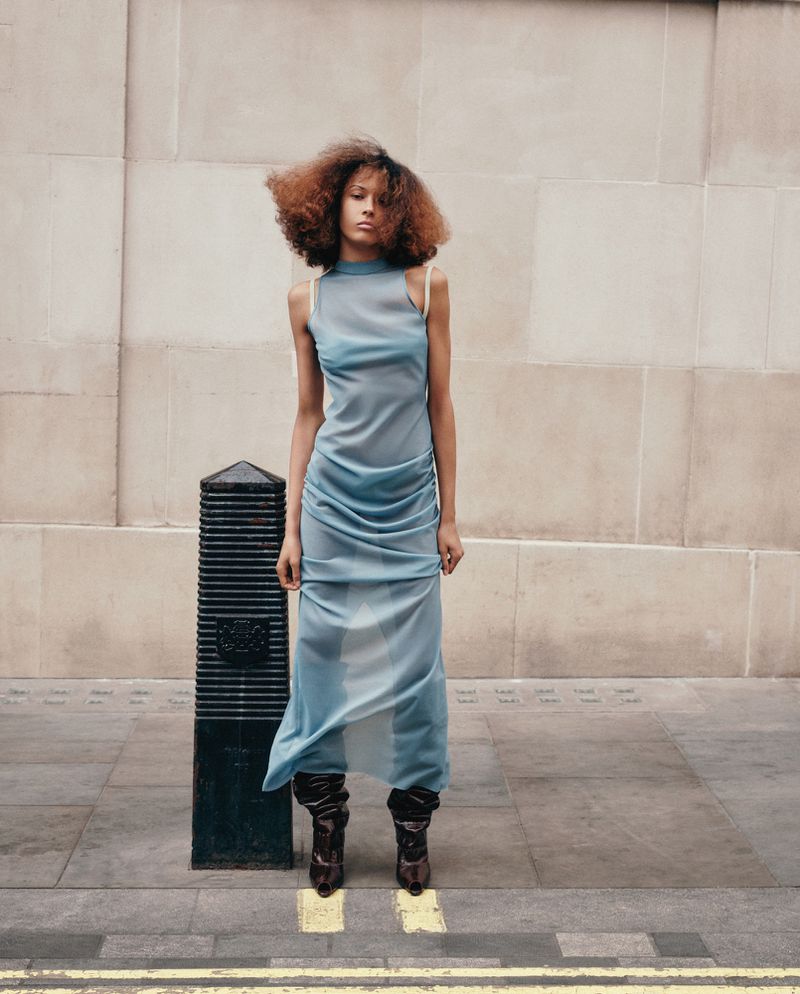 Model Prinnie Stott wearing a sheer powder blue maxi dress and dark slouchy boots standing beside a street bollard.
