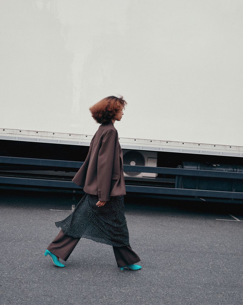 A woman in a brown suit walks on an asphalt surface, with a large white truck in the background, on a bright overcast day.