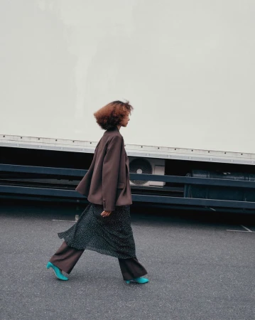 A woman in a brown suit walks on an asphalt surface, with a large white truck in the background, on a bright overcast day.
