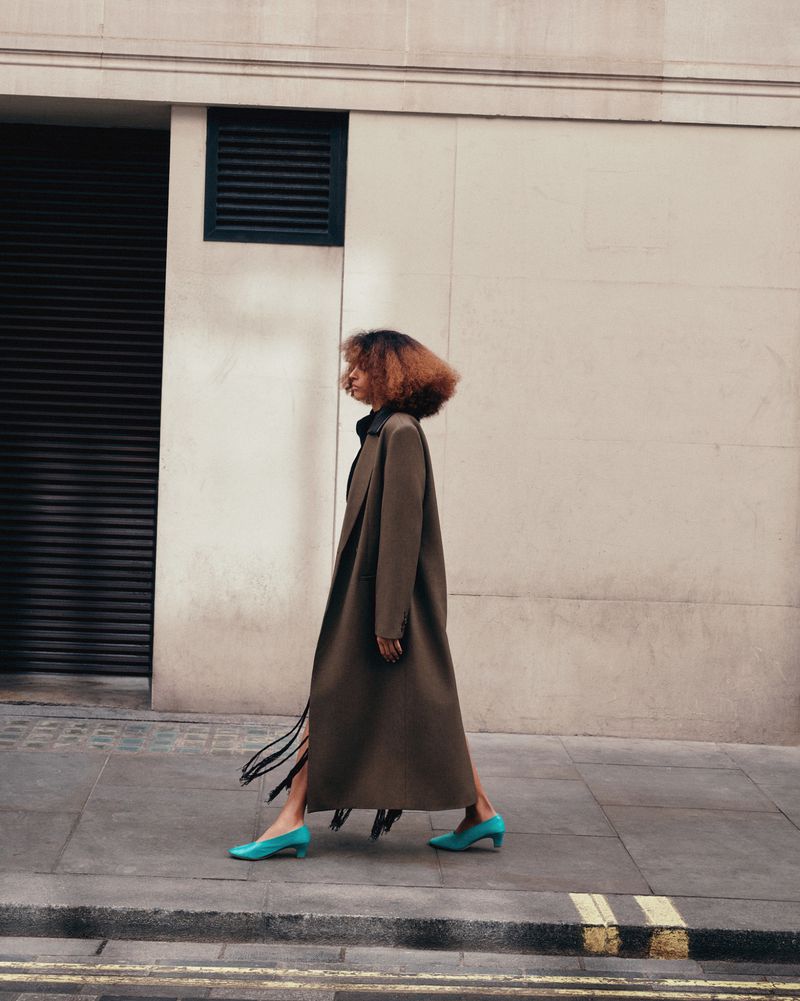 A woman in an olive coat and blue heels walks along a city street, showcasing a full-length fashion shot on a sunny day.