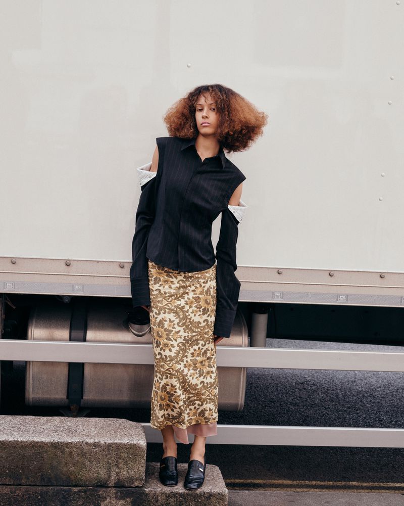 A woman with curly hair poses in a black shirt and printed skirt, standing on stone steps with a white truck in the background.