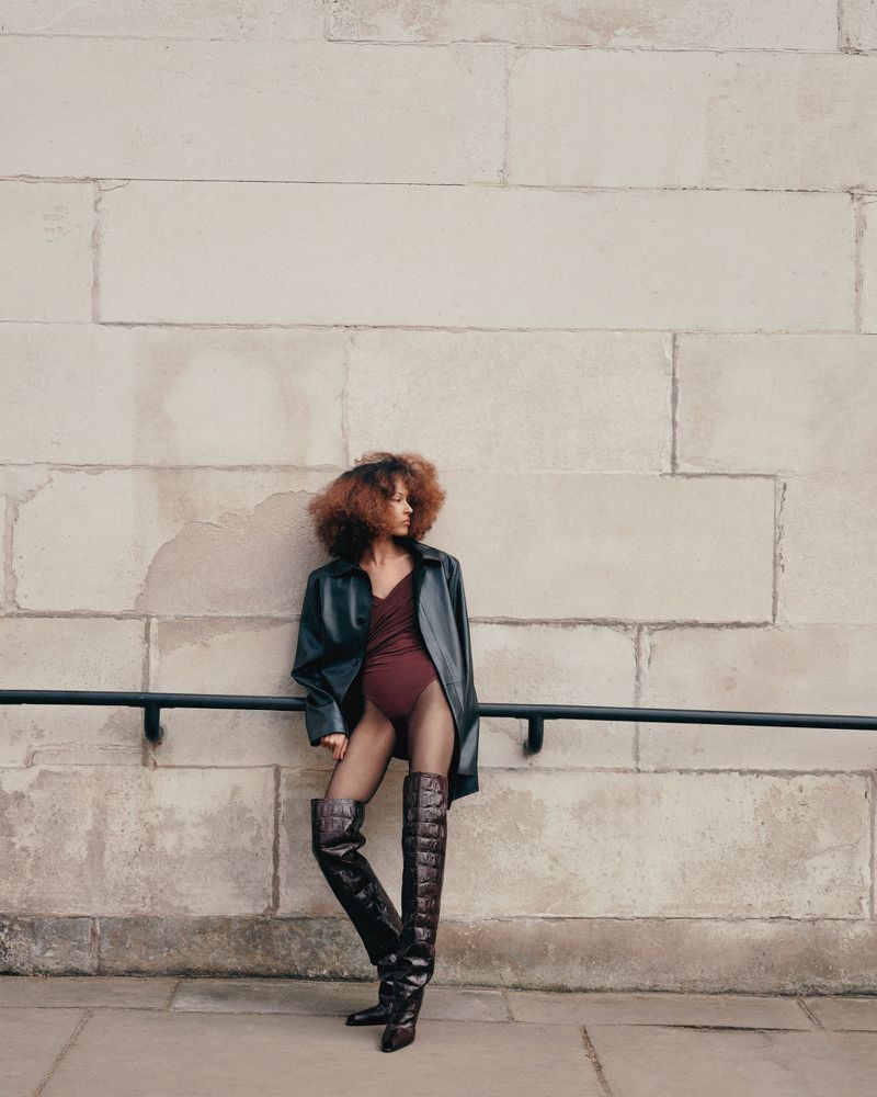 Stylish adult woman poses in a black coat, maroon bodysuit, thigh-high boots, and fishnet stockings in an urban setting.