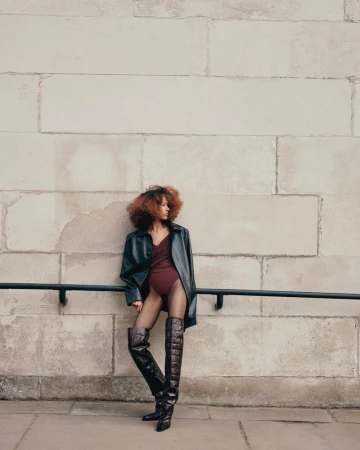 Stylish adult woman poses in a black coat, maroon bodysuit, thigh-high boots, and fishnet stockings in an urban setting.