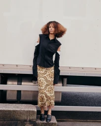 Model Prinnie Stott wearing a black cutout shirt and a patterned gold midi skirt standing in front of a white truck.