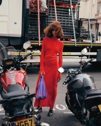 Model wearing a long red dress holding a purple plastic bag while standing between two motorcycles on an urban street.