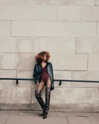 Model Prinnie Stott wearing a burgundy bodysuit, dark jacket, and thigh-high boots leaning against a handrail before a stone wall.