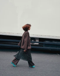 Model Prinnie Stott walking in profile wearing a brown tailored blazer, sheer skirt, and bright turquoise heels beside a white truck.
