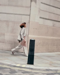 Model Prinnie Stott walking along a city sidewalk wearing an oversized light jacket and color-blocked wide-leg trousers.