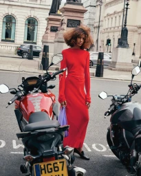 Prinnie Stott wearing a long red dress and black heels standing next to parked motorcycles on a city street.