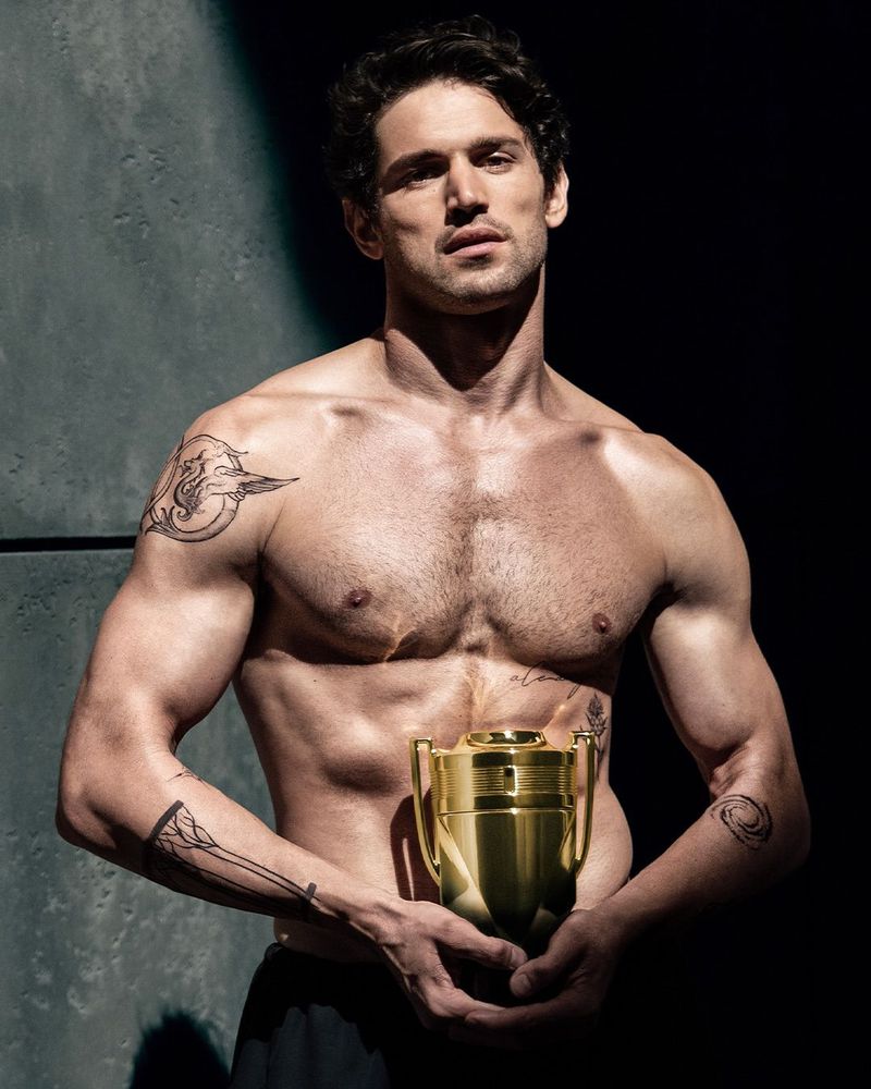 Shirtless man with arm tattoos holding a gold trophy-shaped perfume bottle under dramatic directional lighting.