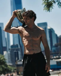Shirtless model Giacomo Cavalli holding a gold trophy on his shoulder against a city skyscraper backdrop.