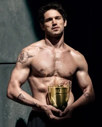 Shirtless man with arm tattoos holding a gold trophy-shaped perfume bottle under dramatic directional lighting.