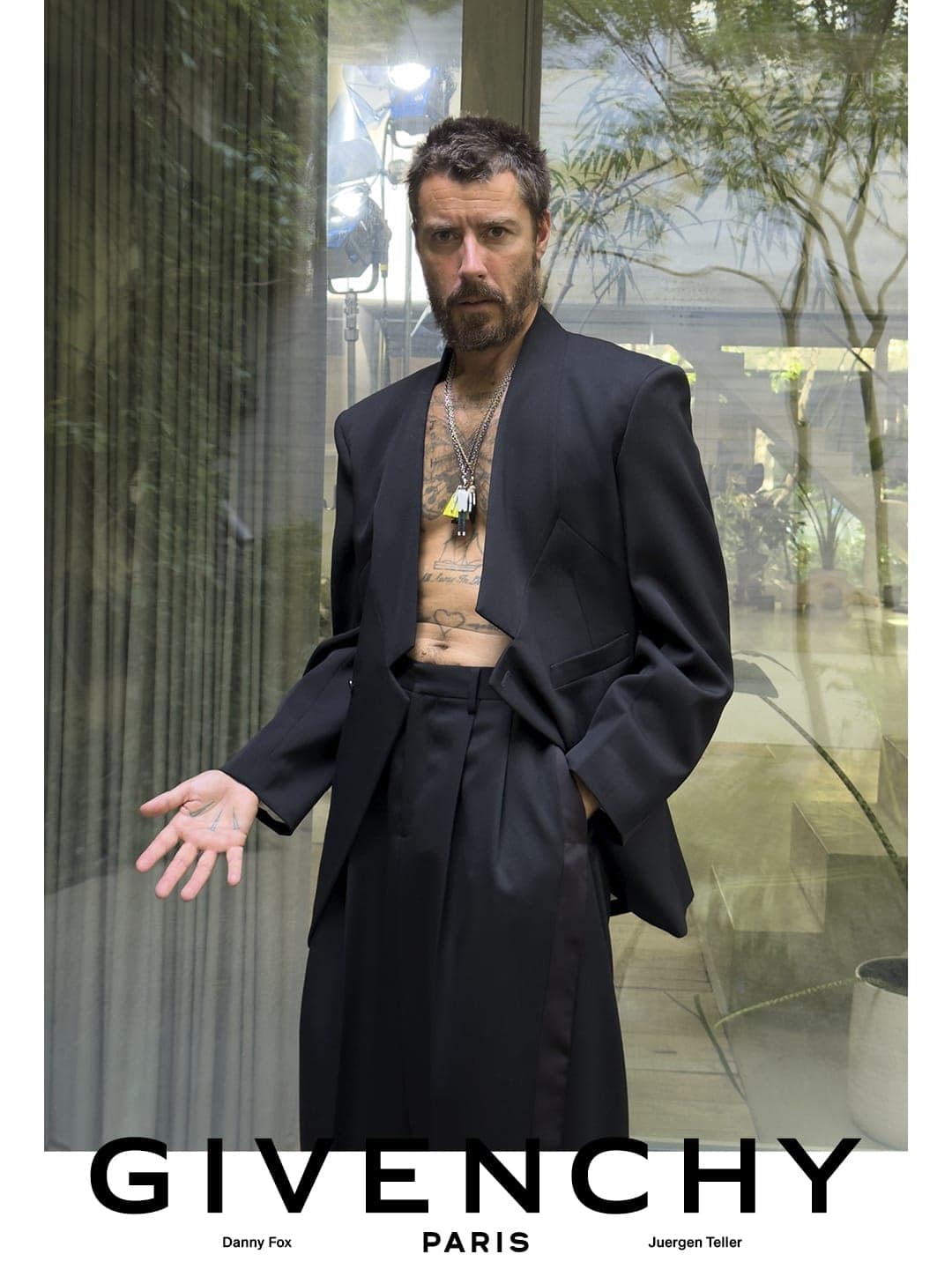 Danny Fox wearing an open black tailored suit with tattoos visible on his bare chest against glass doors.