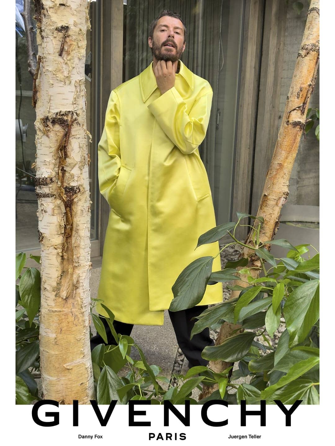 Man wearing a bright yellow coat standing outdoors framed by birch trees for Givenchy menswear campaign.