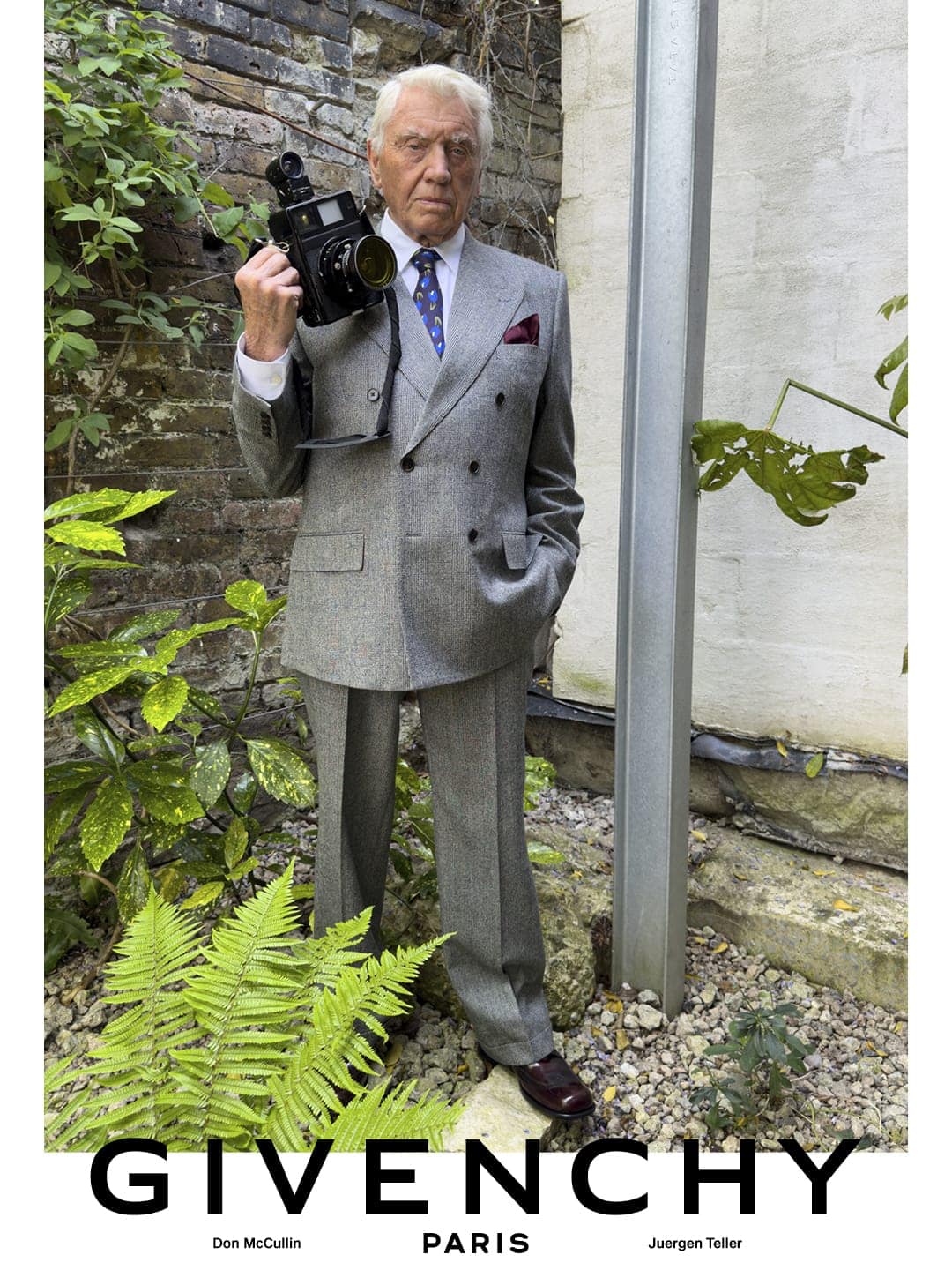 Sir Don McCullin holding a medium format camera wearing a grey double-breasted suit against garden foliage for Givenchy.