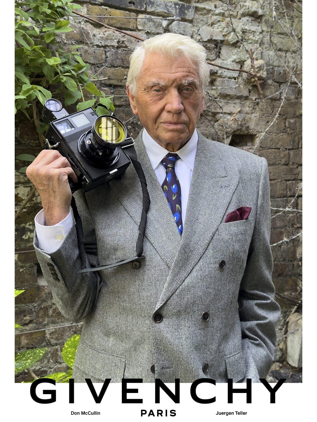 Sir Don McCullin in a grey double-breasted suit holding a vintage camera against a weathered brick wall for Givenchy.