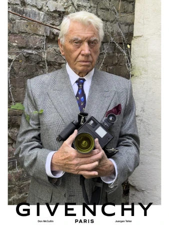 Sir Don McCullin wearing a grey tailored suit and patterned tie while holding a camera in front of a brick wall.