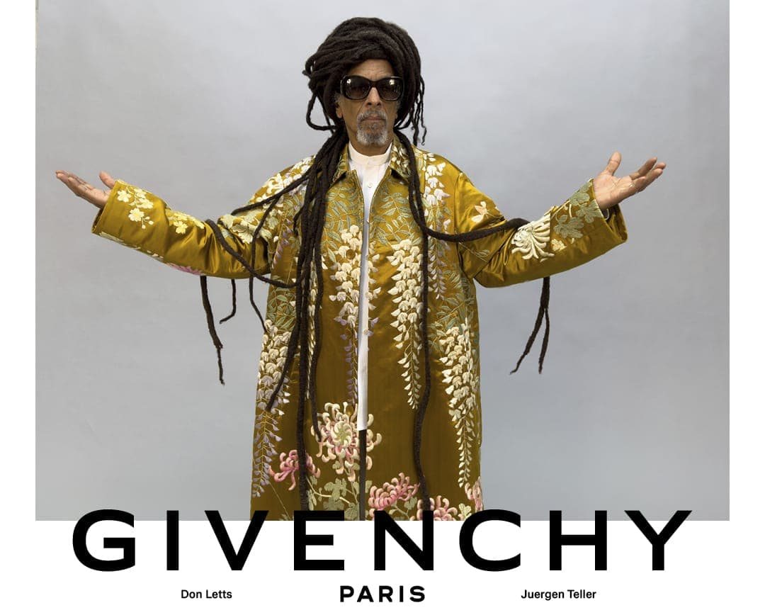 Don Letts poses with arms outstretched wearing an embroidered gold floral coat and sunglasses for Givenchy against a studio backdrop.
