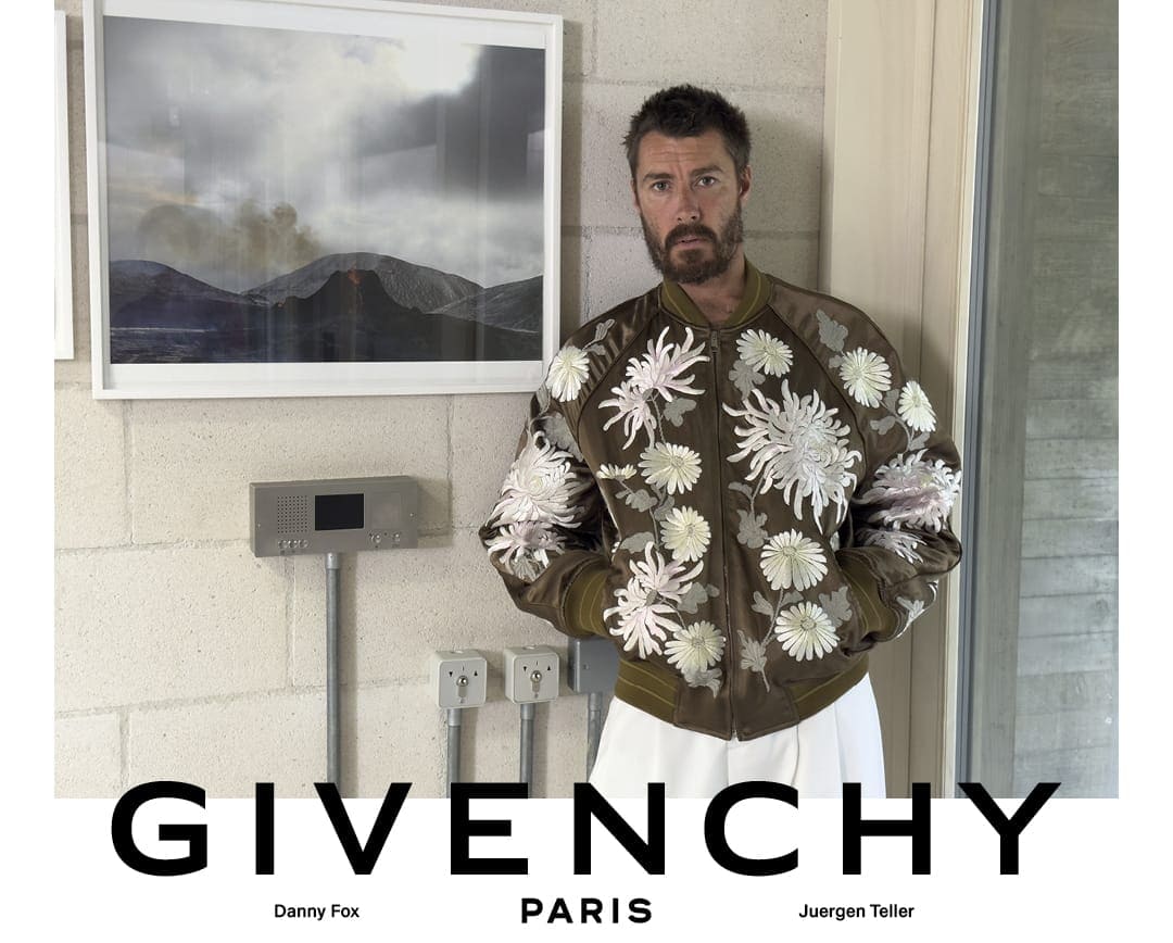 Danny Fox wearing an embroidered floral olive bomber jacket and white trousers against a concrete wall for Givenchy.