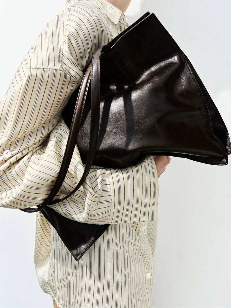 Close-up of a person in a cream striped shirt carrying a large dark espresso leather tote bag tucked under their arm.