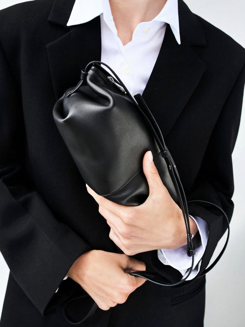 Cropped shot of a model wearing a tailored black blazer and white collared shirt while holding a black leather pouch handbag.