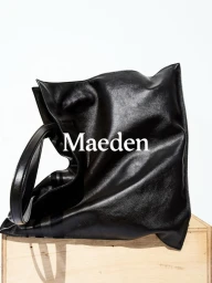 A large black leather tote bag resting on a wooden crate against a clean white background with the Maeden logo.
